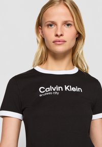 Juodas medvilninis marškinėliai su balta apykakle ir rankovių akcentais. Priekyje yra spausdinimas: "Calvin Klein be galo miesto" balta spalva.