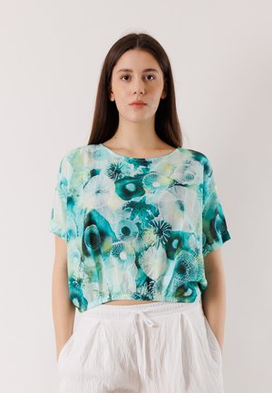 Jonge vrouw met lang bruin haar, gekleed in een tealkleurige en witte bloemetjesprint blouse met korte mouwen en witte broek met trekkoord.