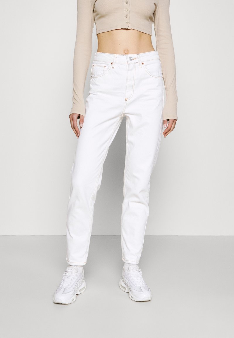 Bílé džíny s vysokým pasem a rovnými nohavicemi, s viditelným prošitím a designem pěti kapes, doplněné bílými teniskami.