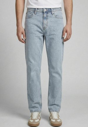 Personne portant un jean bleu clair droit et des baskets blanches avec des accents beige, debout devant un fond gris uni.