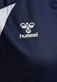 Hvid hummel-logo med stiliseret bi og tekst på mørkeblå stof, del af et sportstøj med hvide og marineblå paneler.