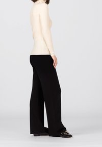 Frau steht seitlich und trägt einen cremefarbenen gerippten Rollkragenpullover, schwarze weite Hosen und schwarze Loafers mit goldener Kettendetail.