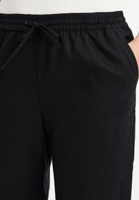 Schwarze Hose mit einem elastischen Bund, ausgestattet mit einem Kordelzug, Seiten Taschen und einer glatten Textur. Der Stoff wirkt leicht und lässig.