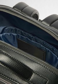 Interno di uno zaino in pelle nera che mostra una superficie texture, una cerniera argentata e un rivestimento blu navy con una piccola etichetta nera sulla tasca interna.