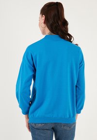 Sudadera azul con cuello y puños acanalados, con un corte holgado y textura suave. Vista desde la parte de atrás, combinada con jeans de mezclilla.