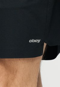 Κοντινή λήψη από το πόδι ενός ατόμου που φοράει μαύρο σορτς με ένα μικρό γκρι λογότυπο «obey» στο κάτω τελείωμα.