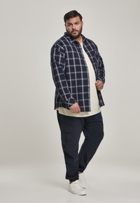 Navy plaid shirt with button closure, worn over a cream t-shirt, paired with navy cargo pants and white sneakers. Smooth fabric texture.
