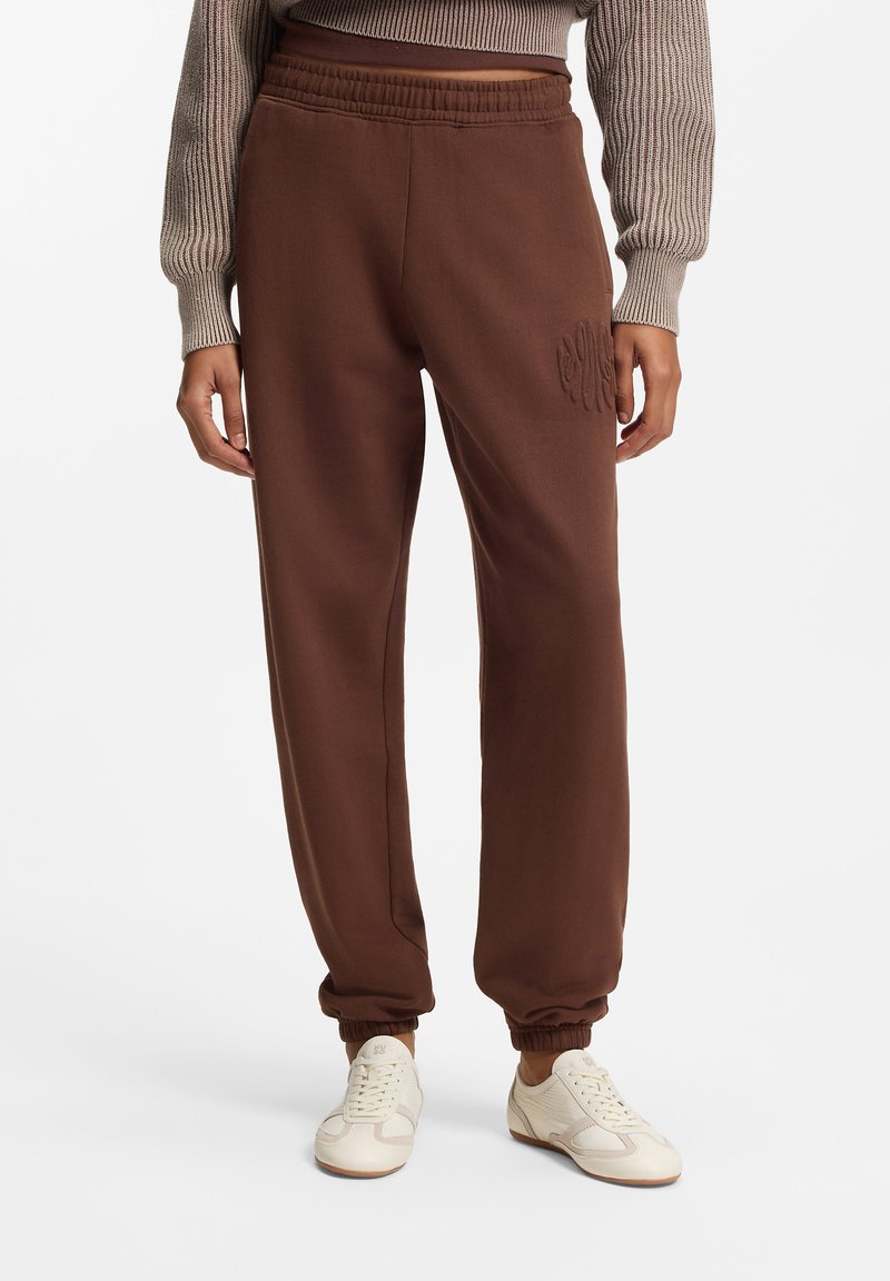 Brune joggingbukser med elastisk talje og manchetter, med et tonalt broderet logodetalje på låret. Glat tekstur, afslappet pasform.