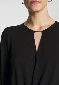 Haut noir avec un décolleté en V et un détail en trou de serrure, fabriqué en tissu doux. Présente des manches longues et un design minimaliste avec un léger éclat.
