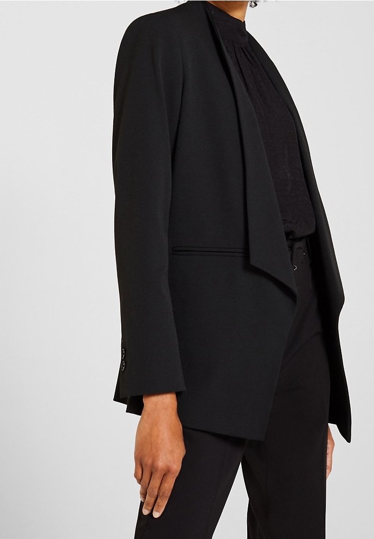 Personne portant un blazer noir sur mesure par-dessus un haut noir et un pantalon noir, debout devant un fond clair uni.