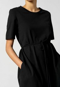 Schwarzes Kleid mit Gürtel, aus weichem Stoff, mit kurzen Ärmeln, rundem Ausschnitt und seitlichen Taschen in einer lockeren Passform.