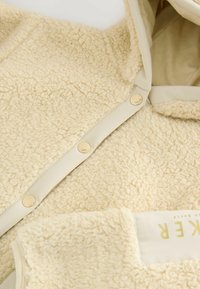 Kermanvärinen fleece-takki, jossa on pehmeä ja teksturoitu pinta, hupullinen, beige-reunus ja kultaiset napit. Piilotettu tasku edessä.