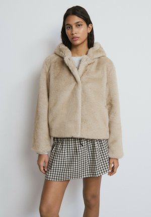 Beige Kunstpelz-Cropped-Jacke mit Kapuze, die eine weiche Textur und einen vorderen Druckknopfverschluss aufweist. Kombiniert mit einem karierten Minirock.