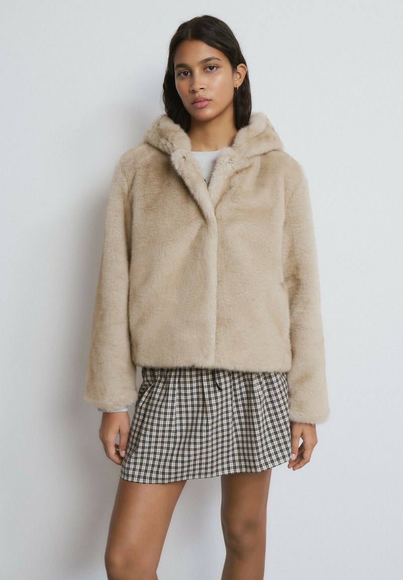 Chaqueta corta de peluche sintético beige con capucha, que presenta una textura suave y cierre frontal con botones de presión. Combinada con una minifalda de cuadros.
