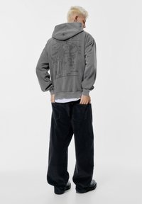 Osoba z krótkimi blond włosami, ubrana w szary polar w kropki na plecach oraz luźne czarne spodnie, stojąca na białym tle.