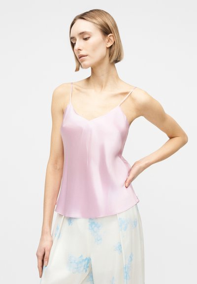 Γυναίκα με κοντά ξανθά μαλλιά που φοράει ανοιχτό ροζ σατέν κορμάκι και άσπρο παντελόνι με μπλε λουλουδάτα σχέδια, σε απλό φόντο.