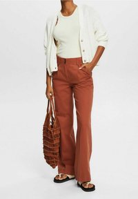 Cardigan blanc à boutons, débardeur crème clair, pantalon large couleur rouille, sac tressé marron, et sandales noires avec accents blancs.