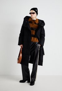 Zwarte gewatteerde jas met een capuchon, oranje en zwart gestreepte zachte trui, zwarte nepleren broek en een bruine ronde handtas.