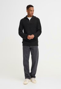 Schwarzer Strickpullover mit hohem Kragen und halbem Reißverschluss, mit geribbelter Textur. Kombiniert mit dunkelgrauen Hosen und beigen Sneakers.