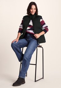 Woman sitting on a tall stool wearing a black sleeveless vest, striped sweater, blue jeans, and black ankle boots, smiling and looking down.