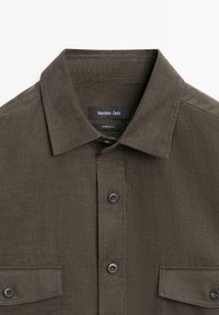 Ciemnobrązowa koszula ze sztruksu z kołnierzem, dwoma zapinanymi na guziki kieszeniami na piersi i czarnymi guzikami z przodu.