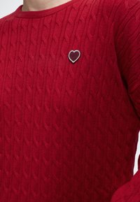 Pull en maille torsadée rouge avec une petite écusson en forme de cœur rouge foncé bordé de blanc sur la poitrine, porté par une personne.