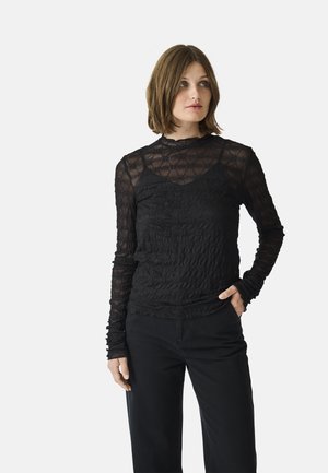 Haut noir à manches longues avec un tissu transparent et texturé présentant des motifs floraux en relief et un col volanté. Associé à un pantalon noir.