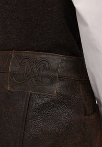 Pantaloni di pelle marrone con una finitura testurizzata, dotati di una toppa in pelle rettangolare impressa con una lettera "M" stilizzata sulla vita.