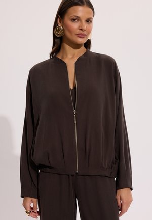 Kobieta w oversizowej ciemnobrązowej kurtce zapinanej na zamek, do której dobrane są spodnie, duże złote kolczyki i złoty pierścionek na lewej ręce.