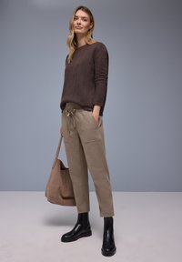 Donkerbruine trui met gestructureerd breisel, beige broek met trekkoord en zwarte enkellaarzen. Houdt een zachte, lichtbruine totebag vast.