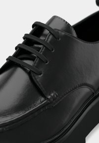 Juodos odinės bateliai su blizgiu paviršiumi, turintys raištelių dizainą, ryškius siūlus ir plokščią padą stabilumui.