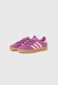 Lila mockaskor med rosa ränder, en strukturerad yta och gummisulor. Har "Gazelle"-logotypen på utsidan.