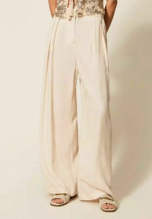 Donna che indossa pantaloni crema larghi a vita alta e sandali avorio con dettagli a catena dorata, in piedi davanti a uno sfondo beige.