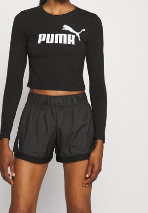 Camiseta corta negra de manga larga Puma con logo blanco combinada con pantalones cortos deportivos negros con cintura elástica en una persona de pie.