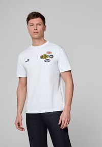 Biała bawełniana koszulka z okrągłym dekoltem, krótkimi rękawami i różnorodnymi kolorowymi naszywkami, w tym flagą Union Jack oraz logiem "WILLIAMS".