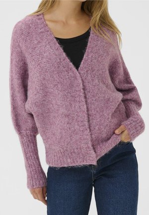 Femme portant un cardigan doux couleur lavande sur un top noir avec un jean bleu foncé, une main dans la poche, sur un fond clair uni.