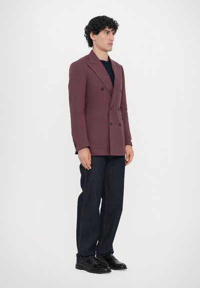 Doppelreiher in kastanienbraun mit strukturiertem Schnitt, kombiniert mit dunkelblauen Jeans und schwarzen Schuhen. Glattes Material mit subtiler Schimmer.