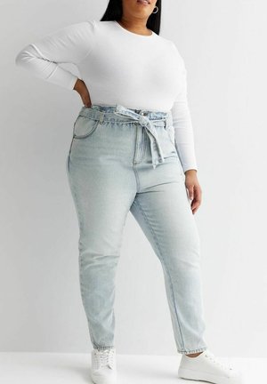 Kobieta ubrana w jasnoniebieskie jeansy z wysokim stanem i przewiązanym paskiem, białą bluzkę z długim rękawem oraz białe trampki, stojąca na jednolitym tle.