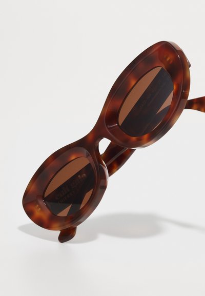 Lunettes en écaille de tortue avec des verres ovales et des montures épaisses. Le design présente des bords arrondis et une finition brillante. Les verres bruns filtrent la lumière du soleil.