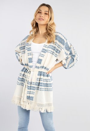 Vrouw met lang golvend blond haar, die een witte top, lichtblauwe spijkerbroek en een wit-blauwe kimono-stijl cardigan met franjes draagt.