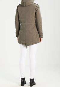 Personne portant une parka à capuche taupe, un pantalon blanc ajusté et des bottines noires, debout de dos.