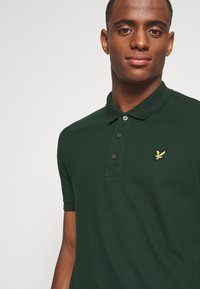 Mørkegrønn polo skjorte med krage, tre svarte knapper, og en gul brodert logo på venstre bryst. Korte ermer, strukturert stoff.
