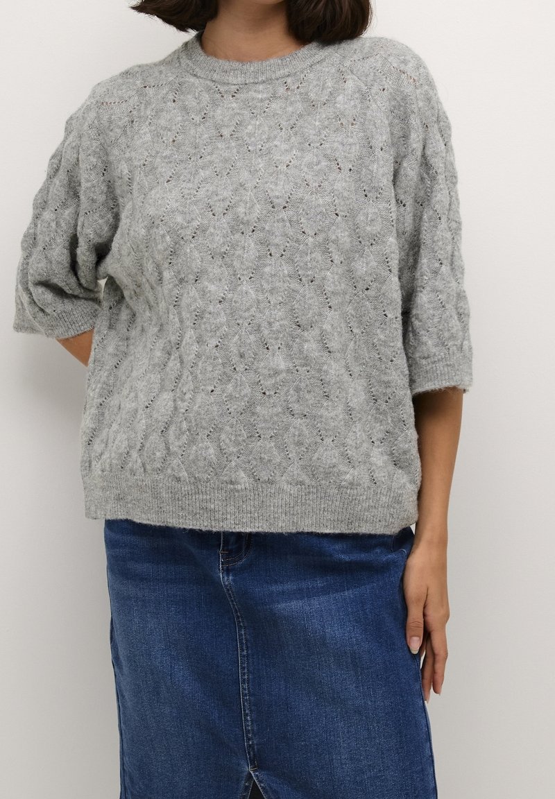 Femme portant un pull en maille torsadée gris clair avec des manches longues jusqu'au coude et une jupe en jean bleue sur un fond uni.