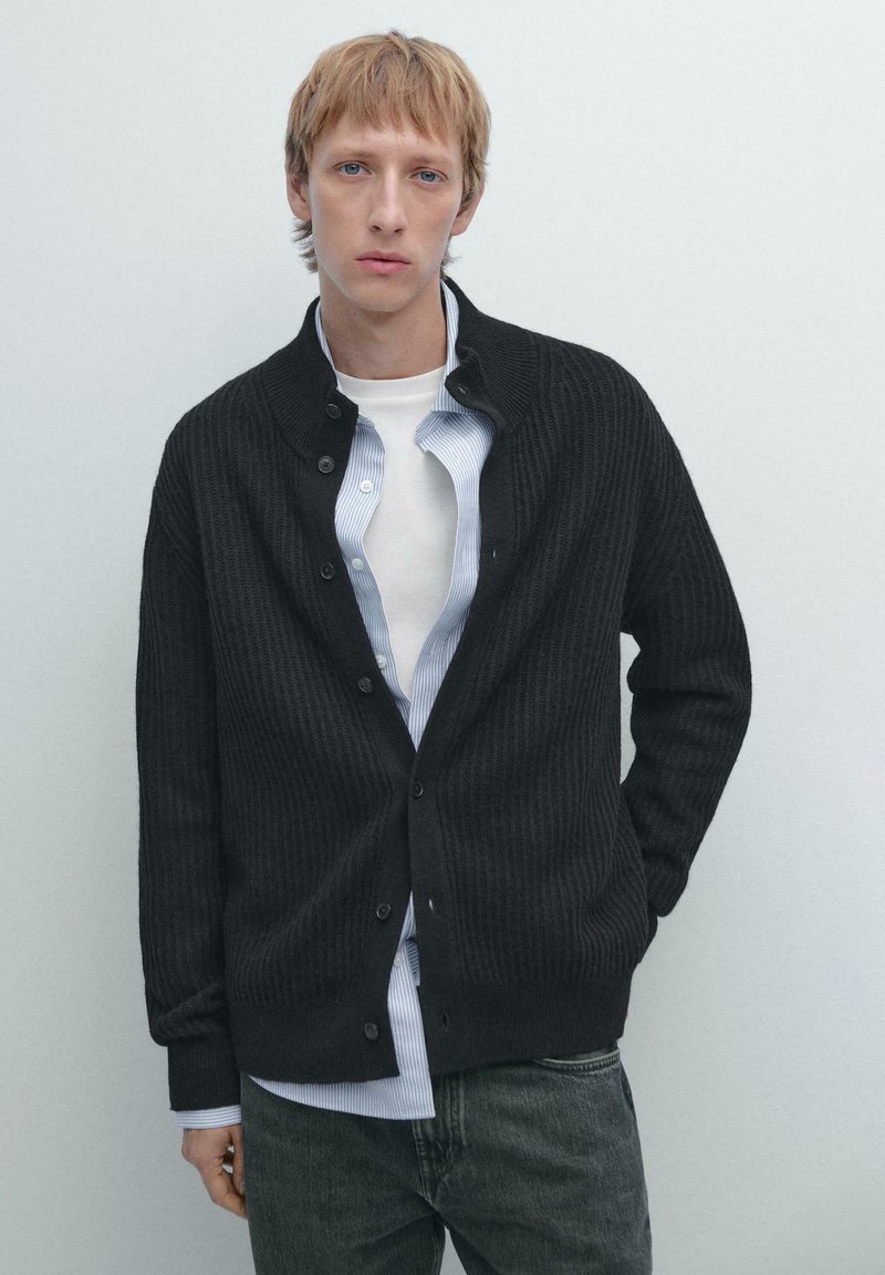 Dark ribbed cardigan made of knitted fabric, button-up design, worn over a light striped shirt, paired with dark jeans.