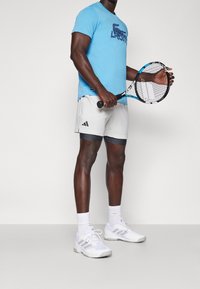 Male tennis player wearing blue Lacoste shirt, white Adidas shorts, white socks, and sneakers, holding a blue and black tennis racket.