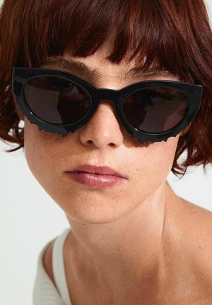 Black cat-eye sunglasses with thick, sculpted frames and dark tinted lenses. Textured edges add distinct, modern detail.