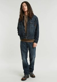 Giubbotto di jeans con due tasche sul petto, colore blu scuro, abbinato a un hoodie marrone e jeans larghi scuri, completato con stivali neri.