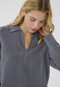 Blouse en lin gris avec un col en V, des manches longues et des poignets à boutons. Dotée d'une coupe décontractée et d'une texture subtile dans le tissu.