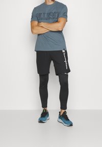 Szary t-shirt z krótkim rękawem z niebieskim logo, czarne szorty z białymi detalami, czarne legginsy pod spodem oraz niebieskie buty sportowe.