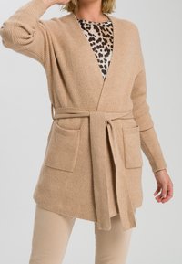 Kvinde iført en beige strikket cardigan, der er bundet om taljen over en leopardmønstret top og beige bukser, med den ene hånd hævet nær sit hoved.